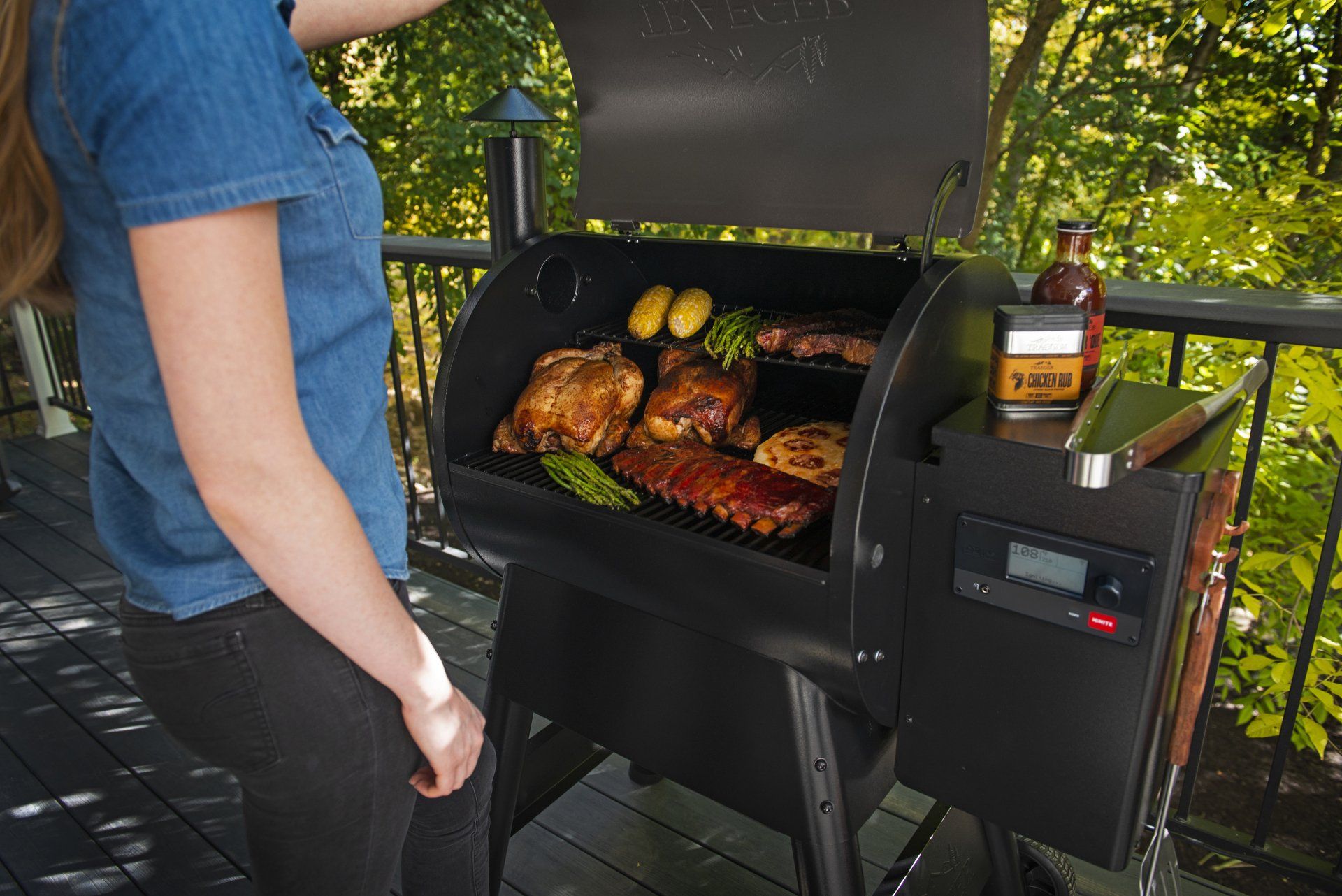 Une personne, debout sur une terrasse, ouvre un barbecue à granulés noirs rempli de poulet, de côtes levées, d'asperges et de pommes de terre en train de cuire.