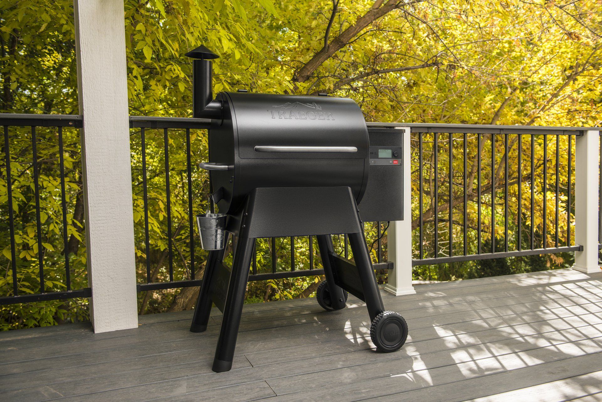 Un barbecue à pellets noir se dresse sur une terrasse en bois, devant une rambarde métallique et des arbres d'automne aux feuilles jaunes.