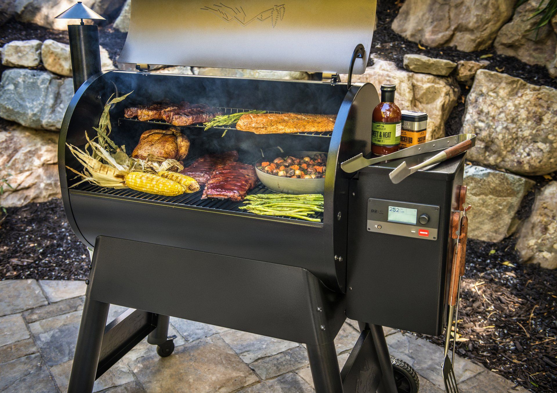 Un barbecue à pellets noirs installé à l'extérieur sur une terrasse en pierre, rempli de viandes, de maïs et de légumes en train de cuire, avec des bouteilles de sauce à proximité.