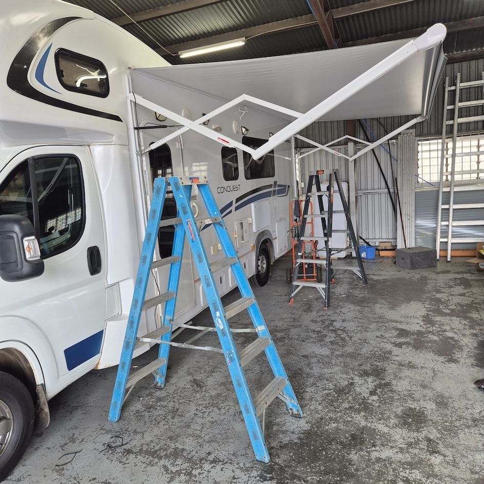 A white campervan with an extended awning. Two ladders stand near the van, inside a garage — Revive Caravan Repairs in Kippa-Ring, QLD