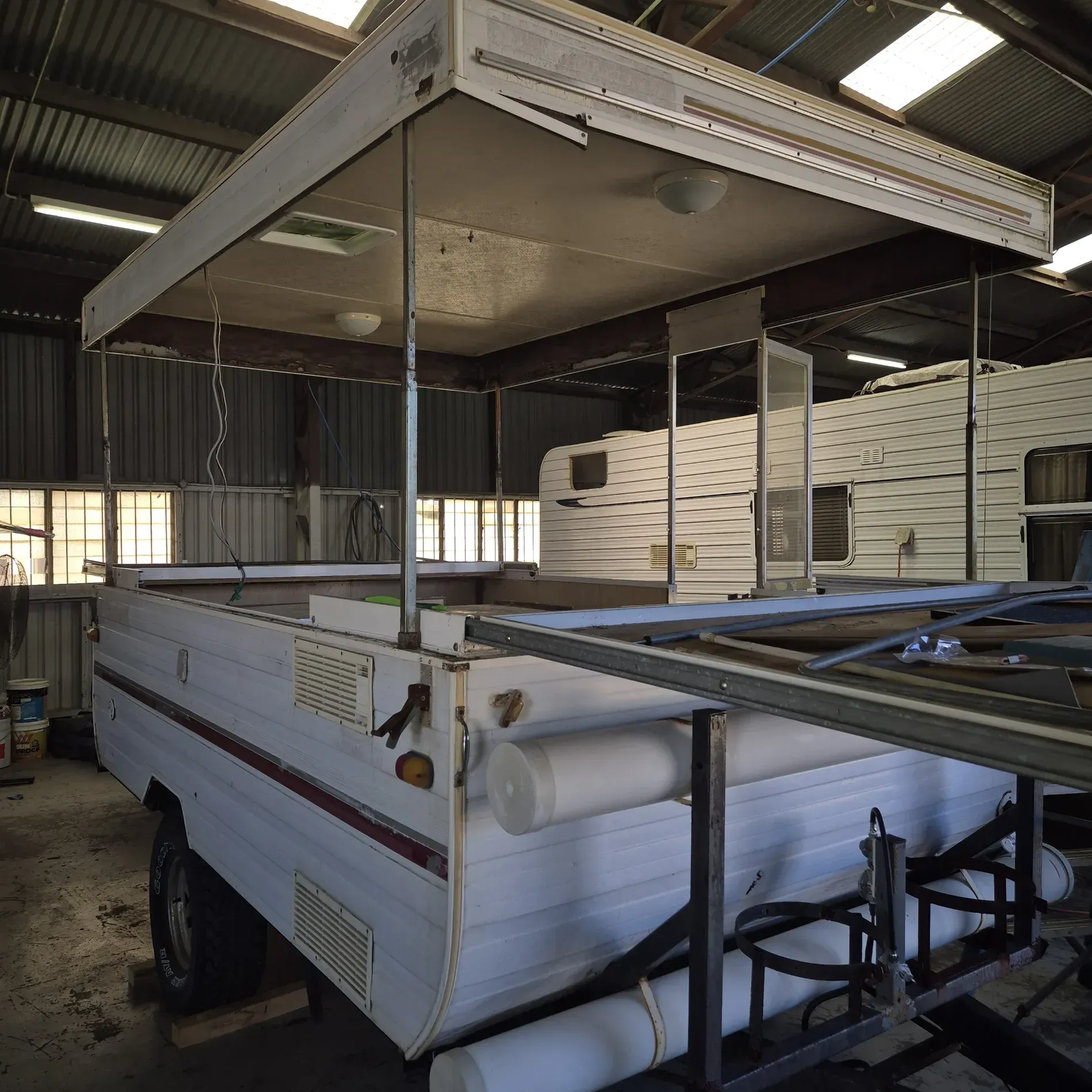 Pop-up camper with roof raised, inside a workshop. White exterior, red stripe, metal framework — Revive Caravan Repairs in Kippa-Ring, QLD