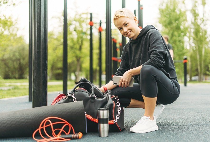 Woman crouching near a gym bag, holding a phone, with workout equipment outside.