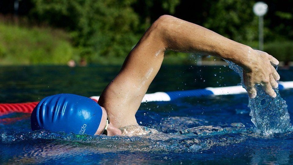 Swimmer in blue cap, arm reaching forward, water splashing, outdoors.