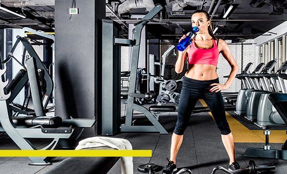 Woman in a gym, drinking from a blue water bottle. She's wearing workout clothes, with weight machines in the background.