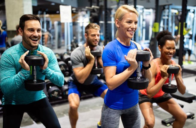 Four people in a gym squat with dumbbells, smiling.