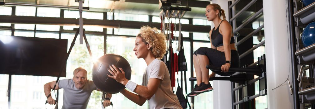 People exercising in a gym: woman holding a medicine ball, another on a machine, and a man using ropes.