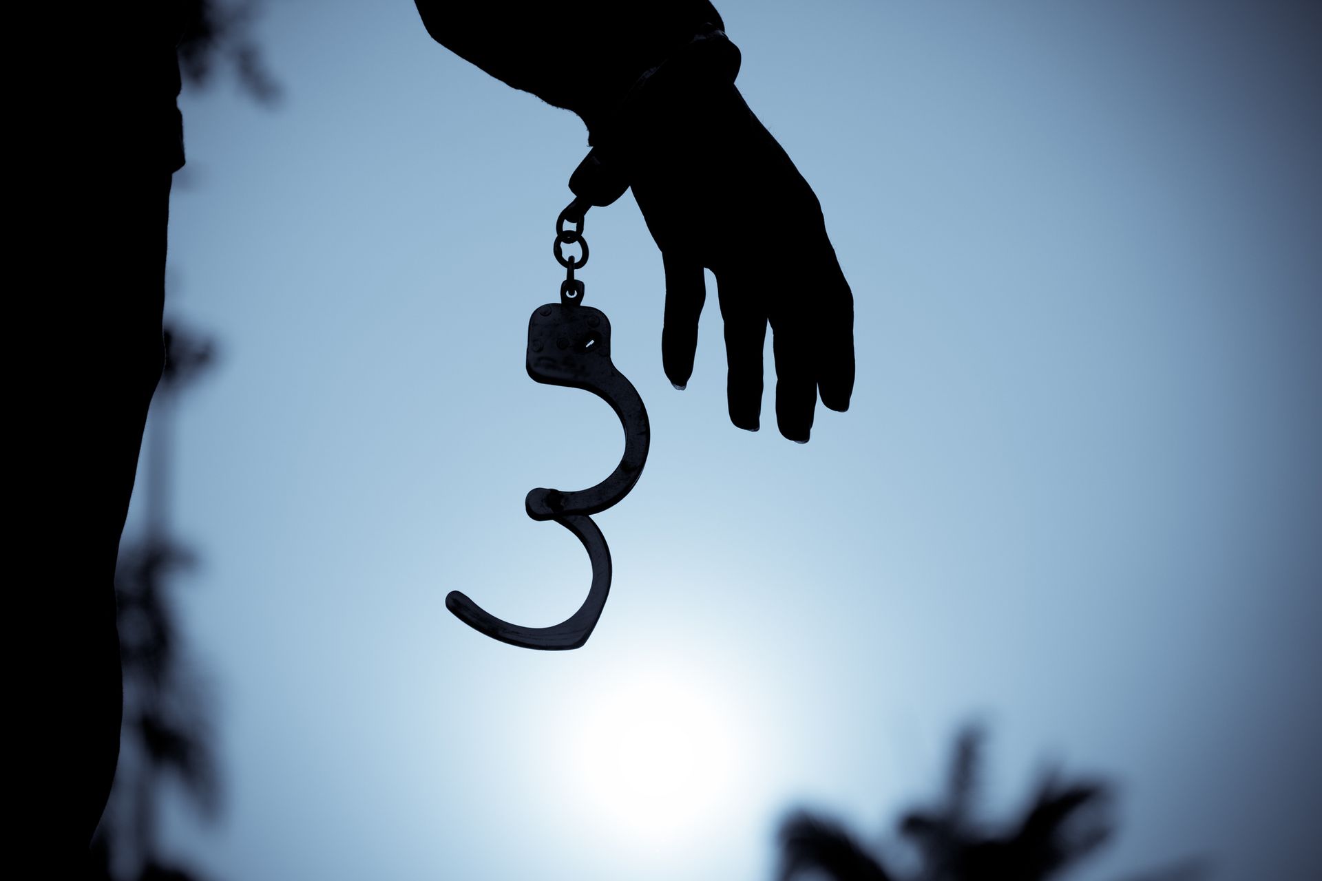 Silhouette of a hand holding open handcuffs against a blue and gray sky.