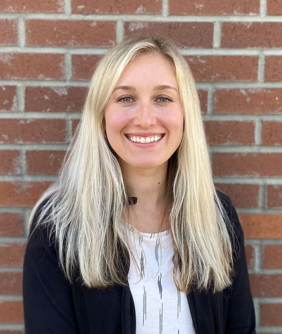 A woman with long blonde hair is smiling in front of a brick wall.