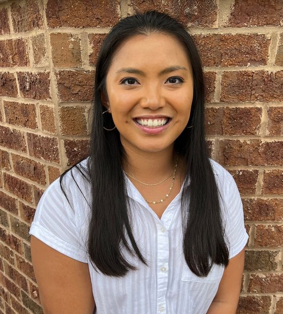 A woman in a white shirt is smiling in front of a brick wall.