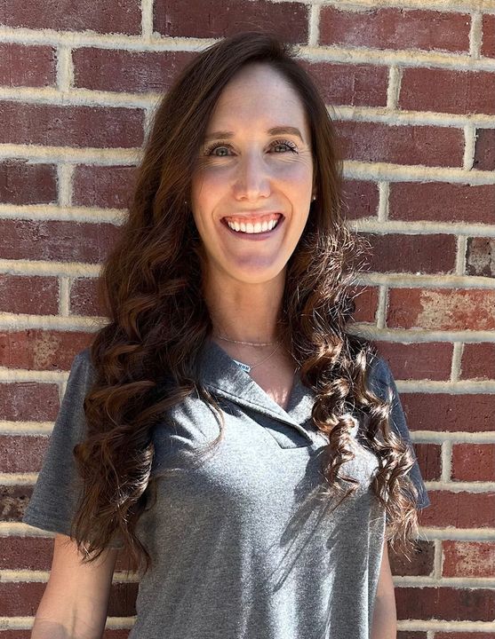 A woman with long red hair is smiling in front of a brick wall.