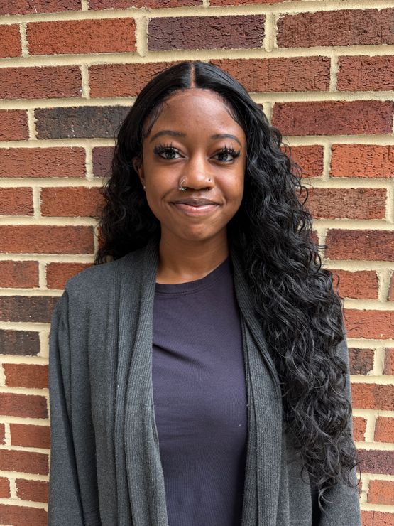 A woman in a black scrub top is smiling in front of a brick wall.