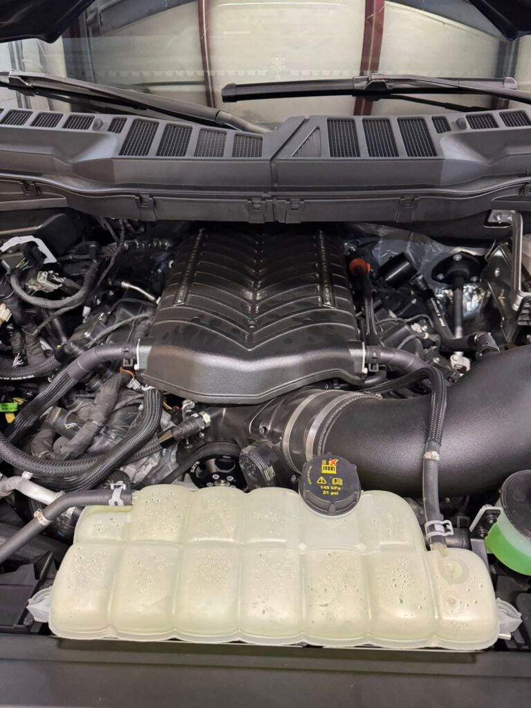 Car engine bay with hood open, showing engine cover, hoses, and coolant reservoir.