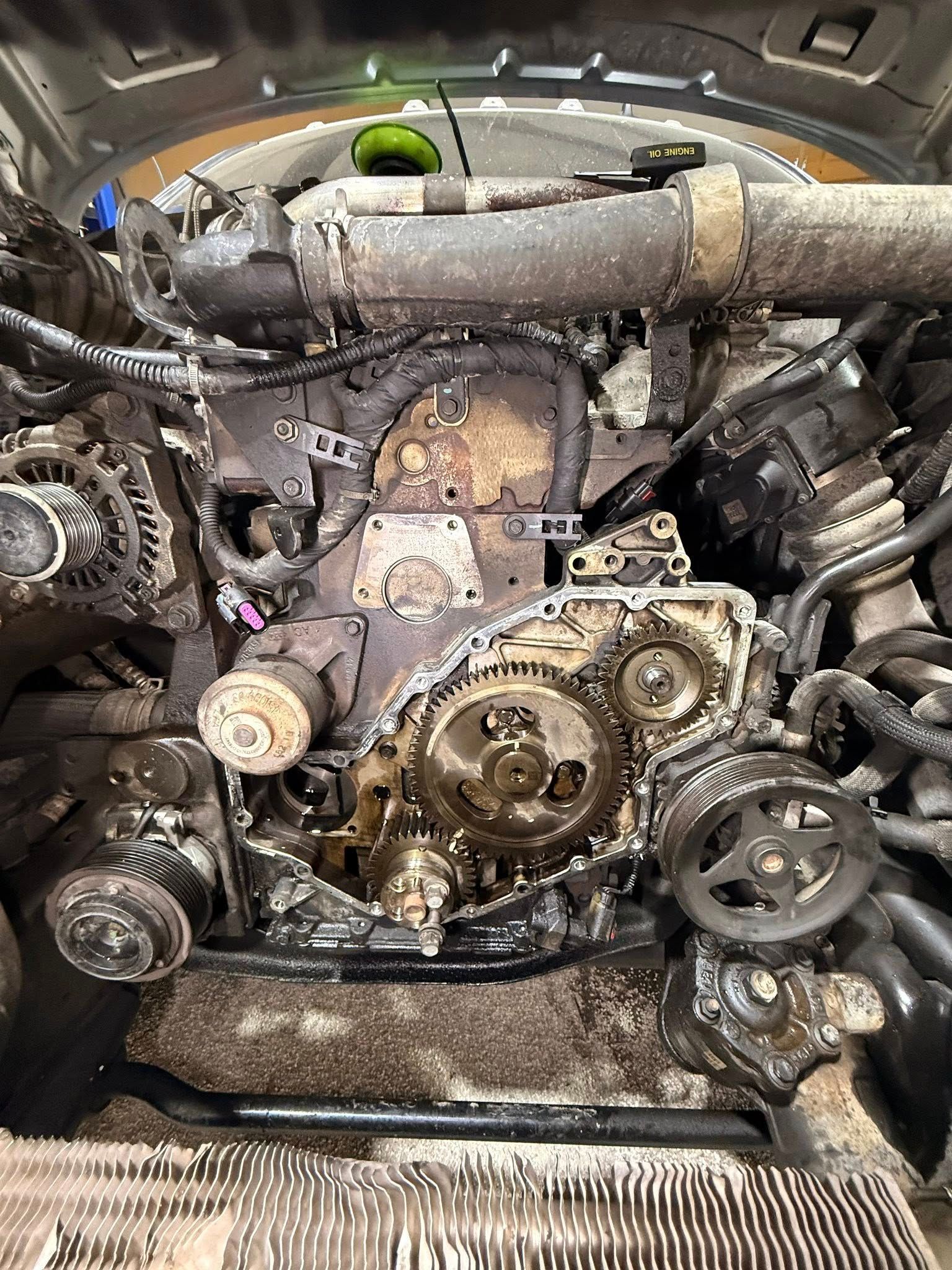 Car engine with front cover removed, exposing timing gears, belts, and internal components.