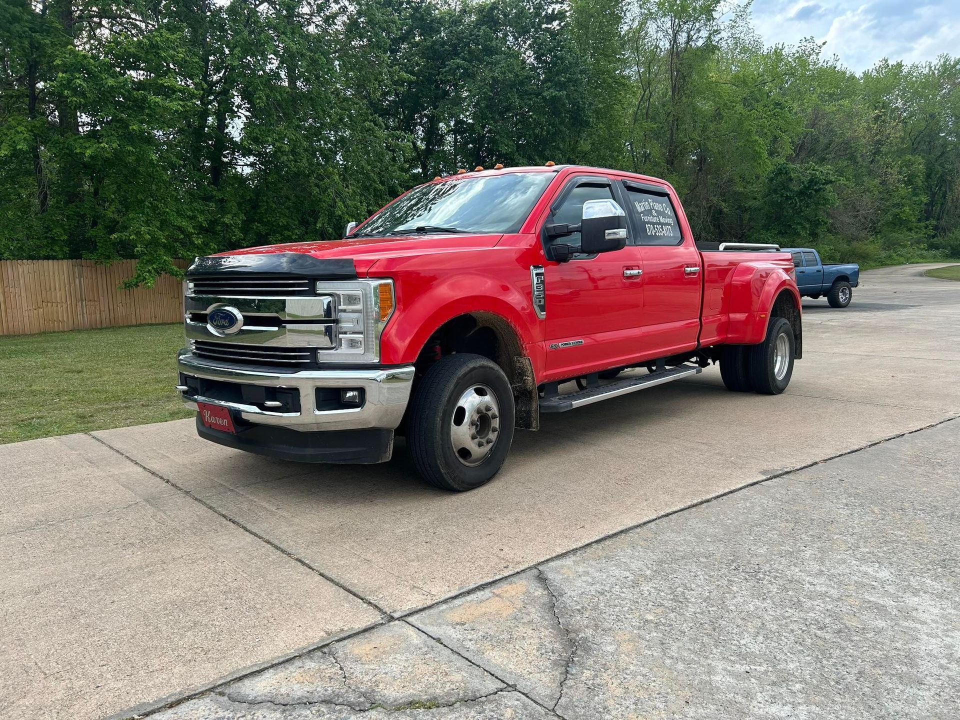 Red Ford pickup truck parked on a concrete lot beside trees and a wooden fence