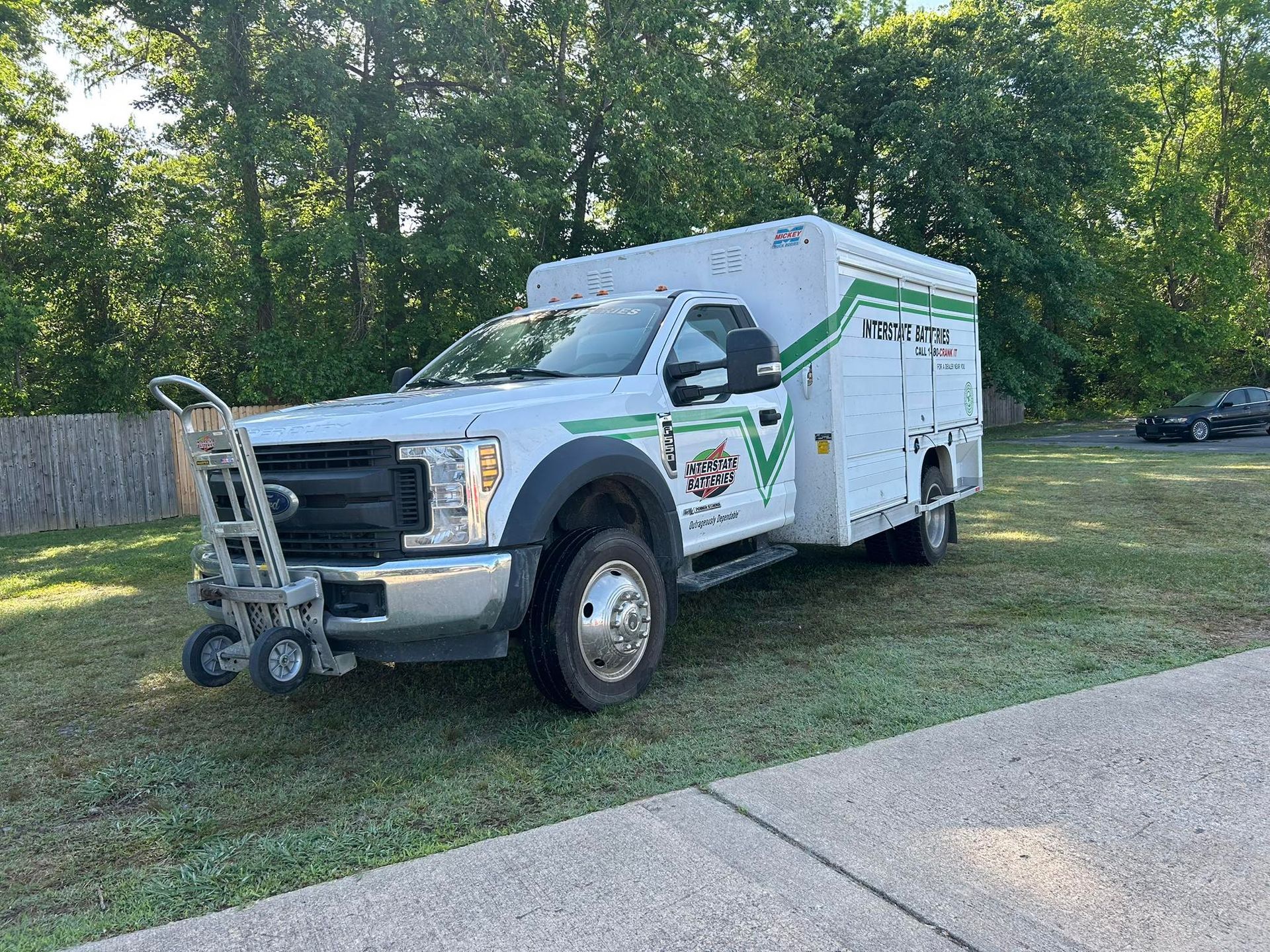 White utility truck parked on grass beside a road, with a ladder rack and trees in the background