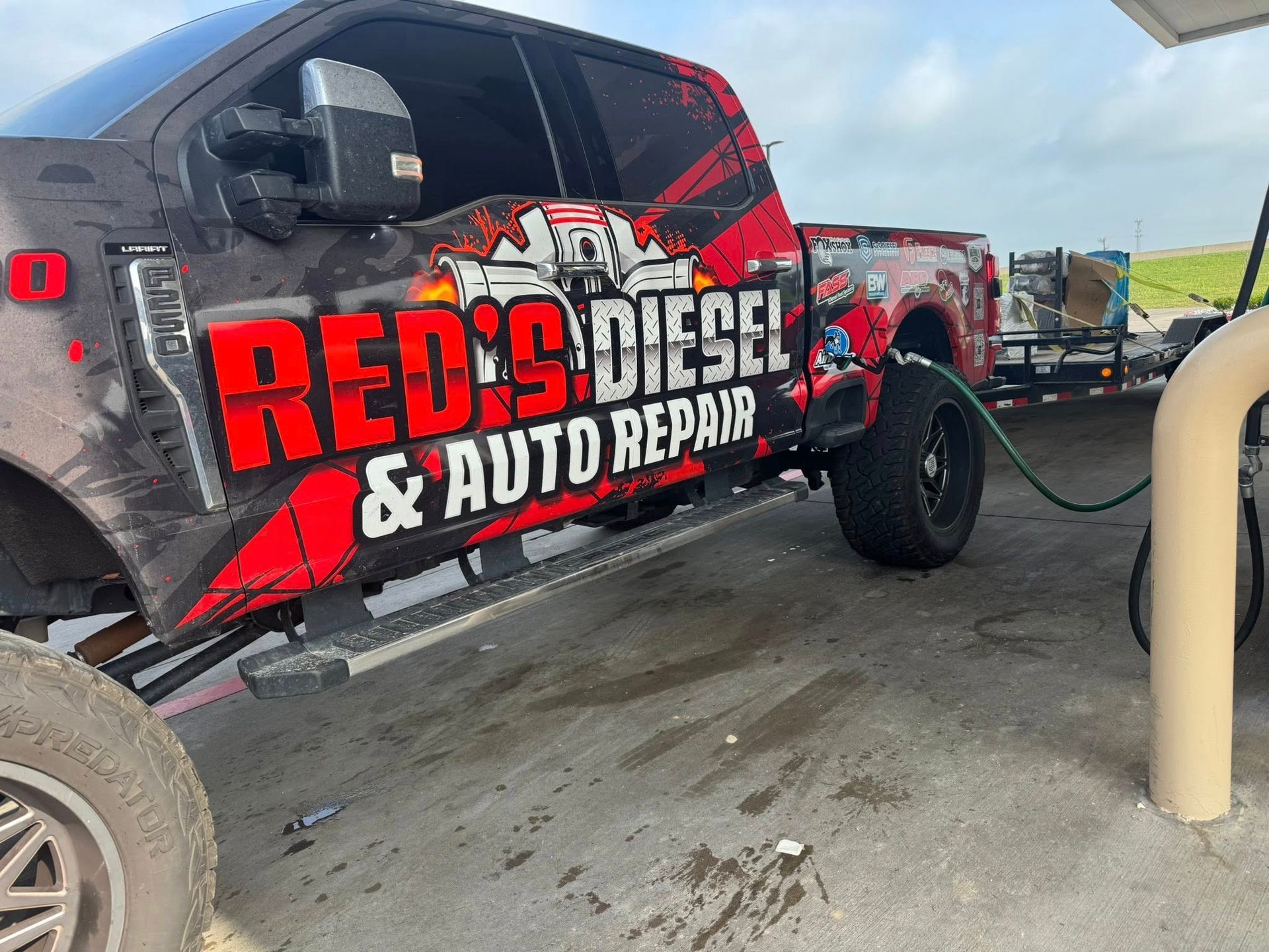 Lifted black truck with red-and-white “RED’S Diesel & Auto Repair” graphics at a gas station pump