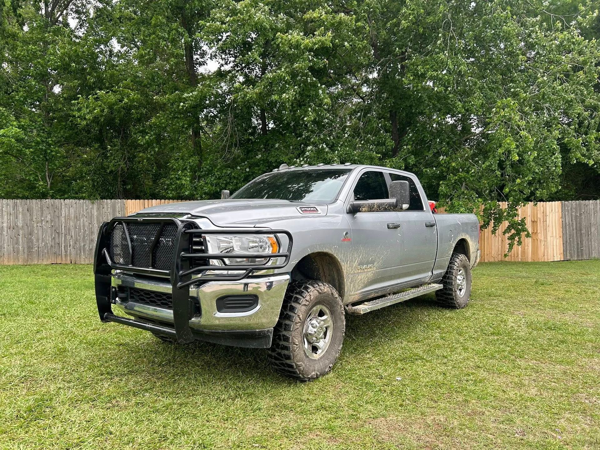 Silver pickup truck with black grille guard parked on grass in front of a wooden fence and trees