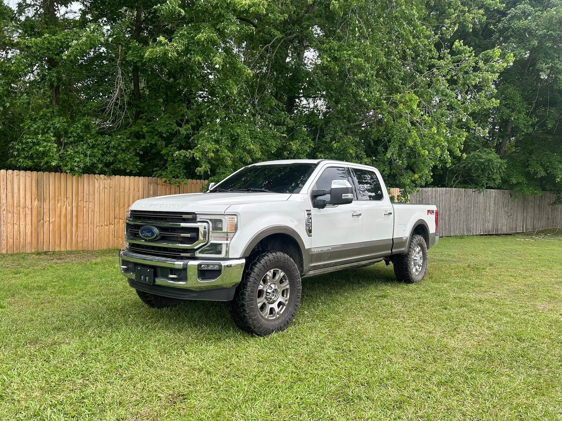 White lifted pickup truck parked on grass beside a wooden fence and trees