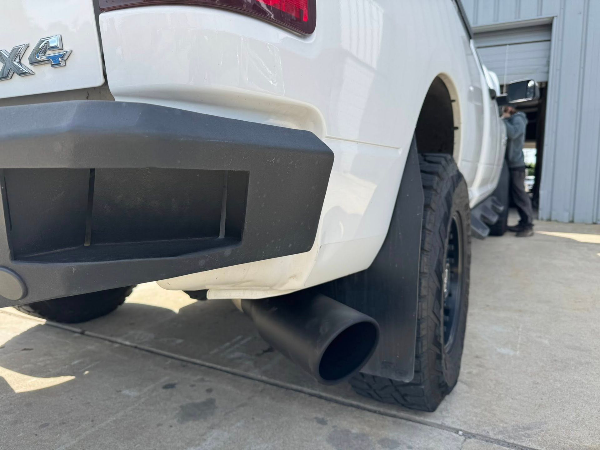 Rear view of a white truck with a large black exhaust pipe, parked beside a metal building.