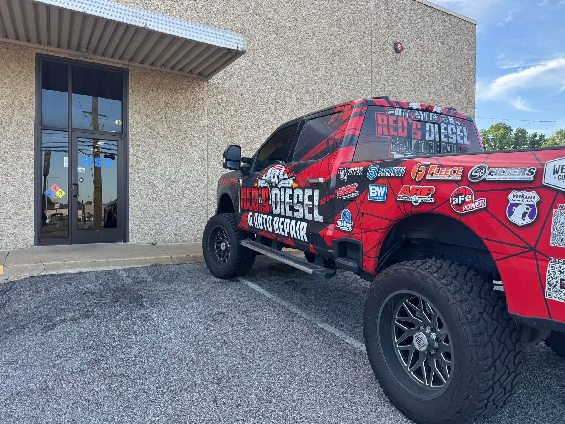 Red off-road race truck parked by a beige building entrance with gravel foreground