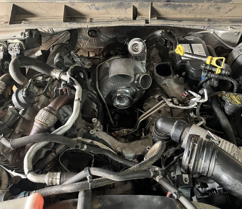 Car engine bay with exposed belts, hoses, and wiring, viewed from above.