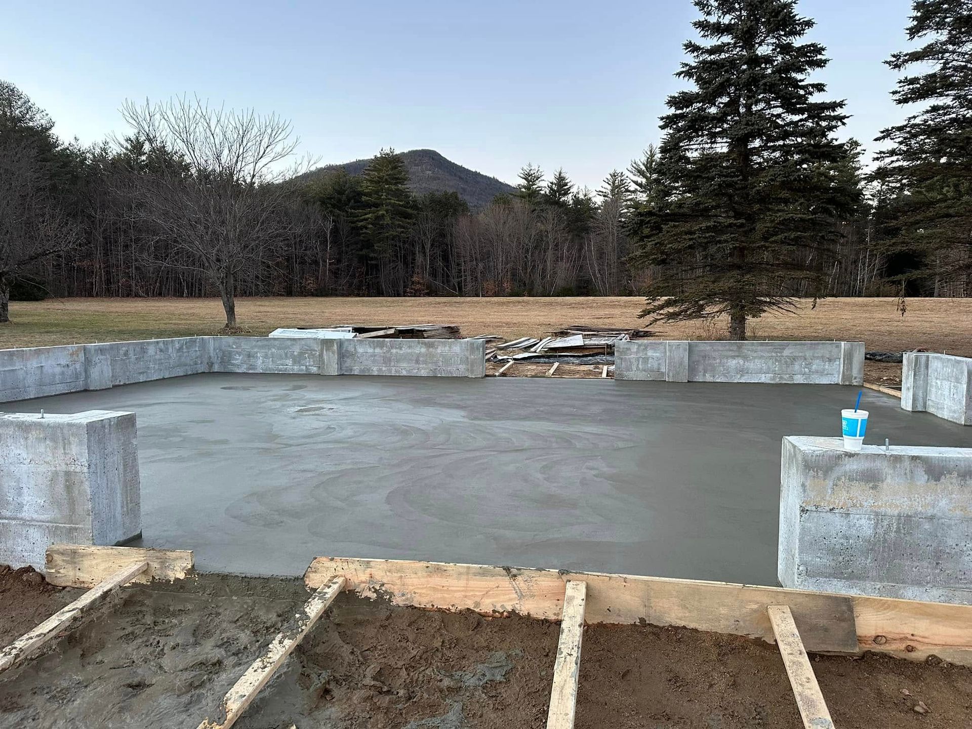 A concrete base for a house is being built in a field with trees in the background.