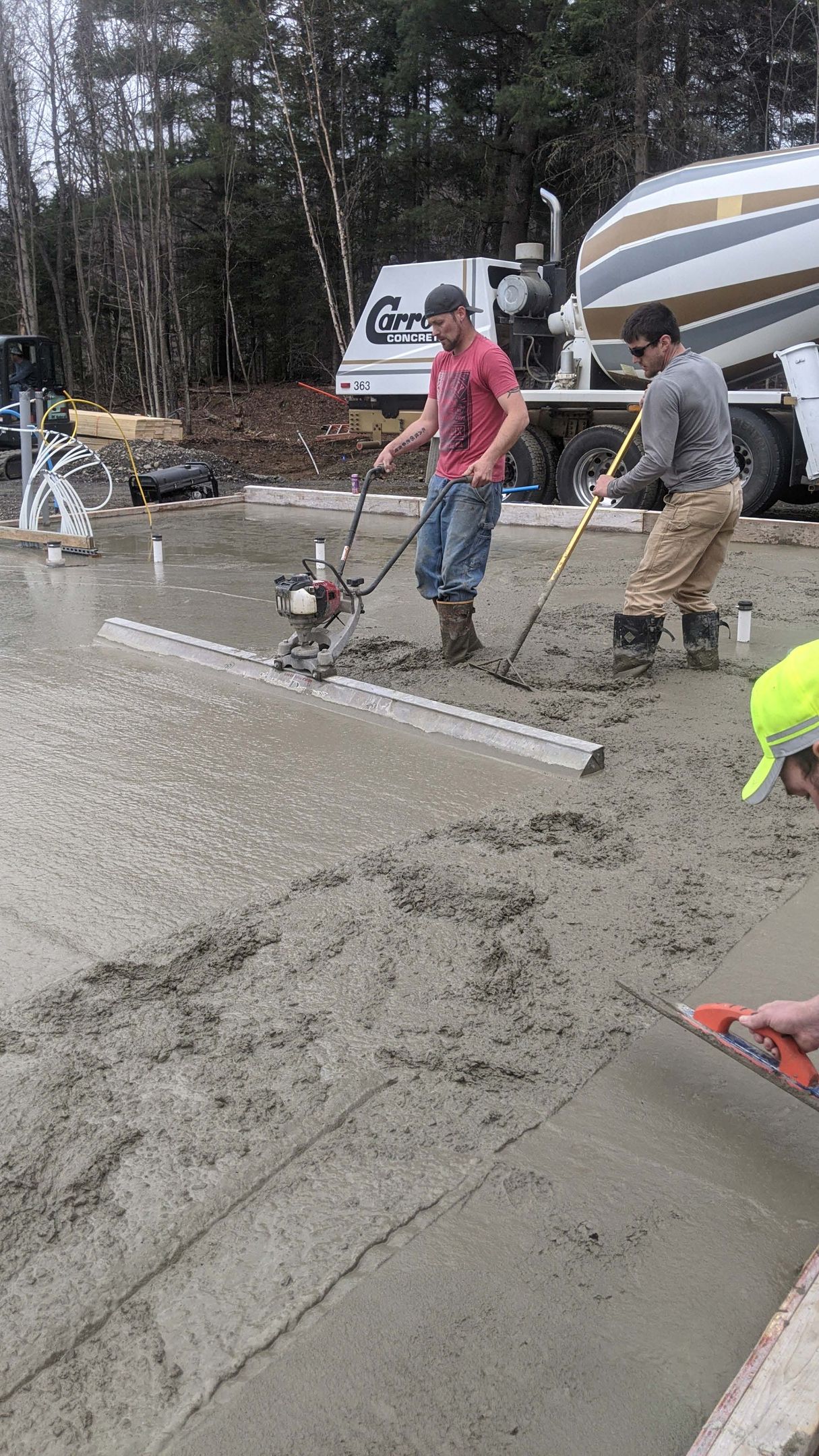 Construction workers smooth wet concrete with a powered screed and hand tools on a residential job site.