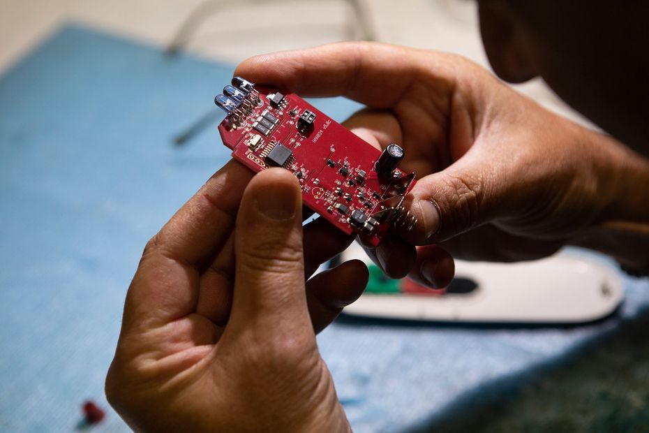 Hands holding a red circuit board over a work surface, with tools blurred in the background