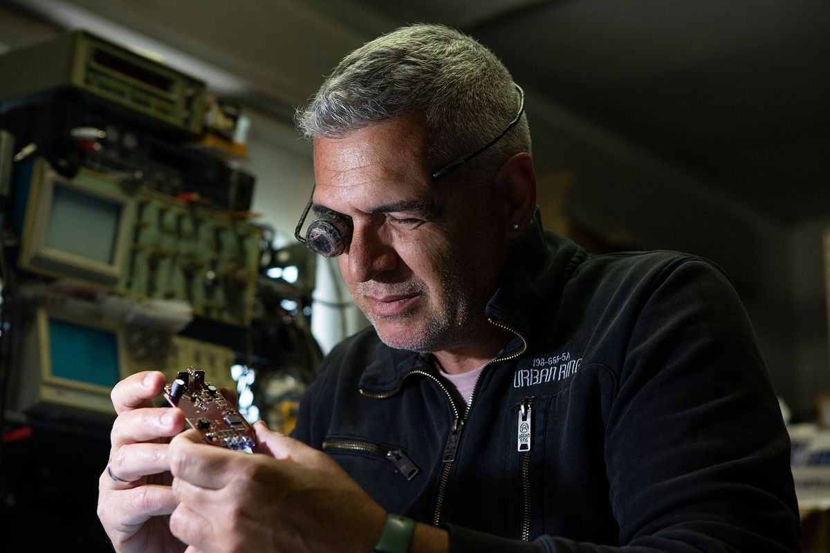 Man examining a small electronic component in a dim workshop, with computer monitors in the background