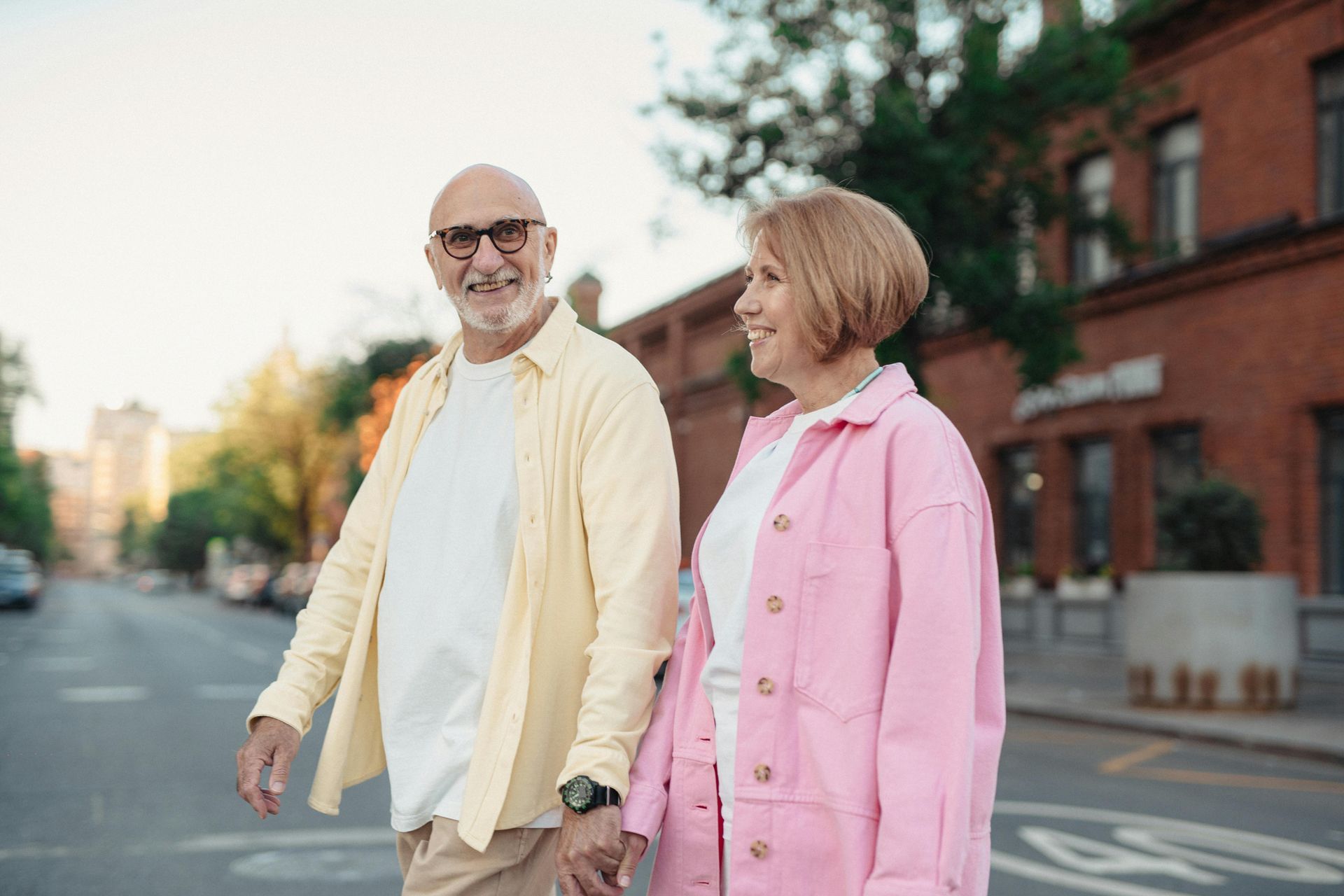 Elderly couple crossing street, smiling. Man in yellow shirt, woman in pink jacket, urban setting.