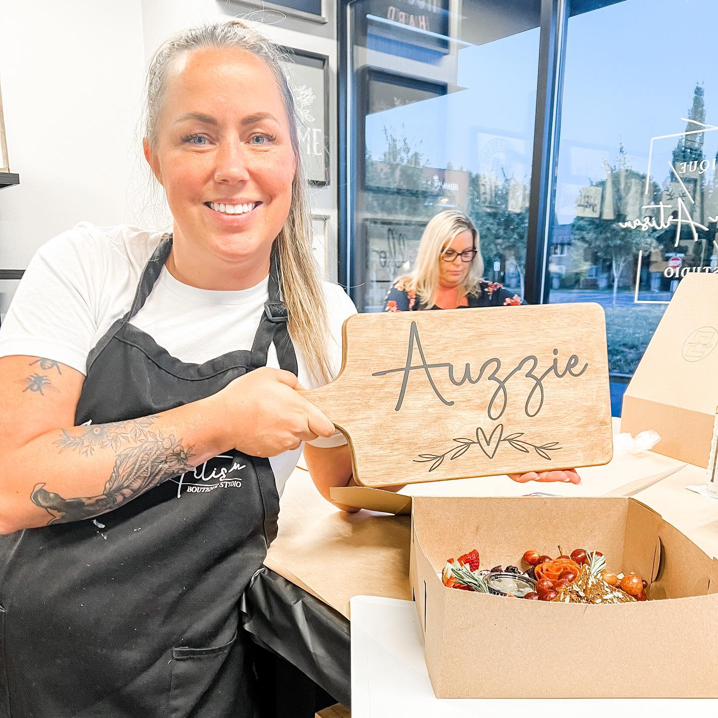 A woman in an apron is holding a wooden cutting board with the name auzzie written on it.