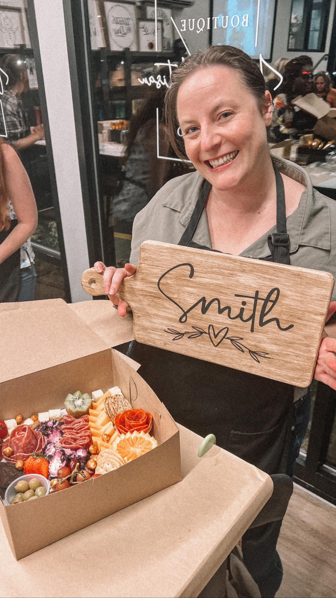 A woman is holding a wooden sign that says `` smith '' next to a box of food.