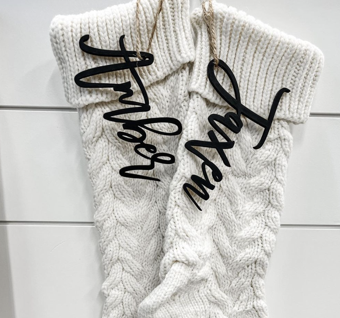 A pair of white knitted christmas stockings with black writing on them hanging on a white wall.