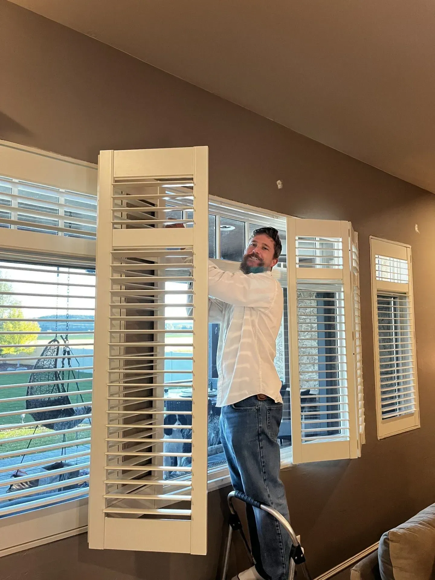 A man is standing on a ladder installing shutters on a window.