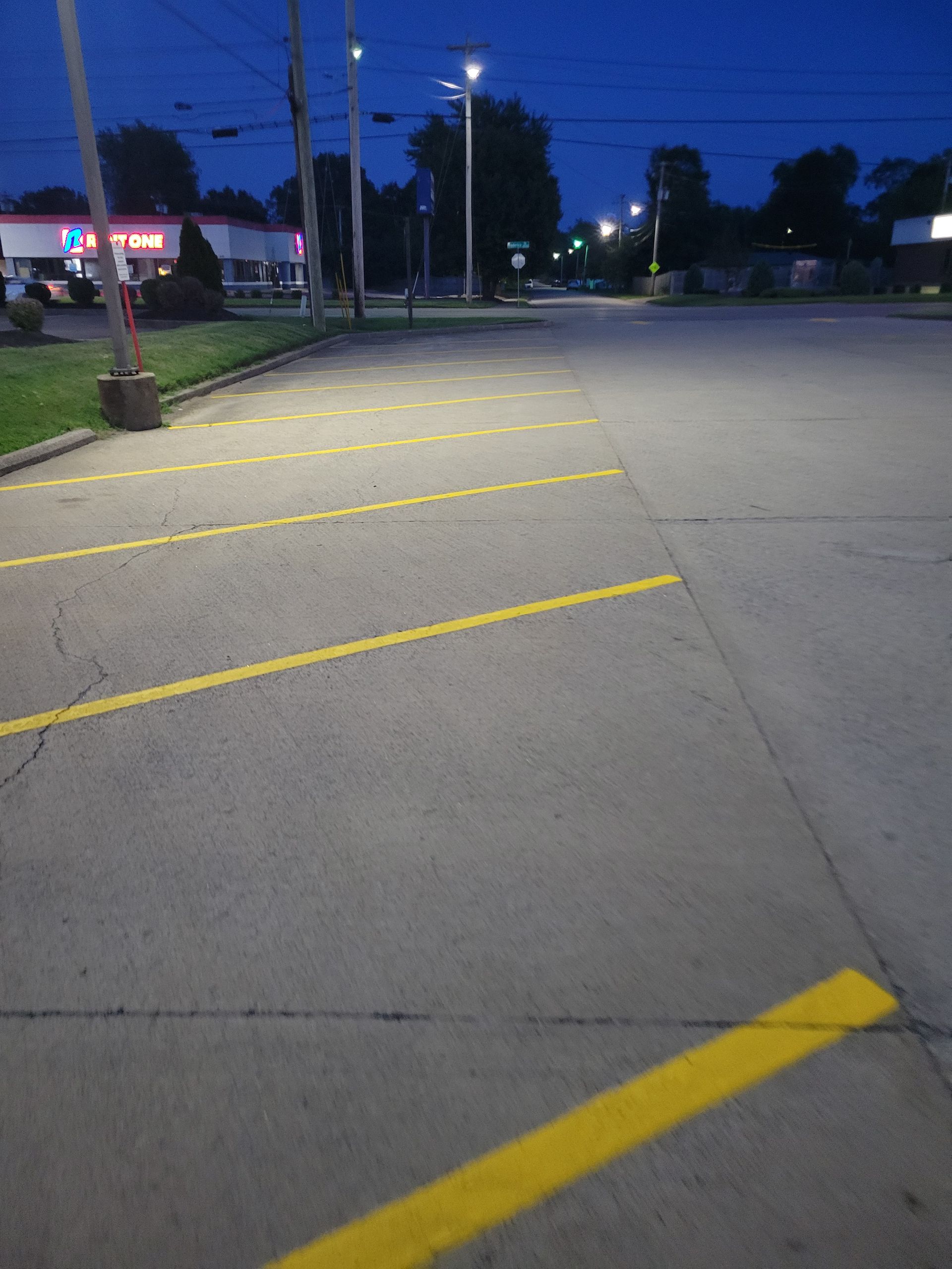 Empty parking lot at dusk; yellow parking lines on gray concrete, lit by streetlights.