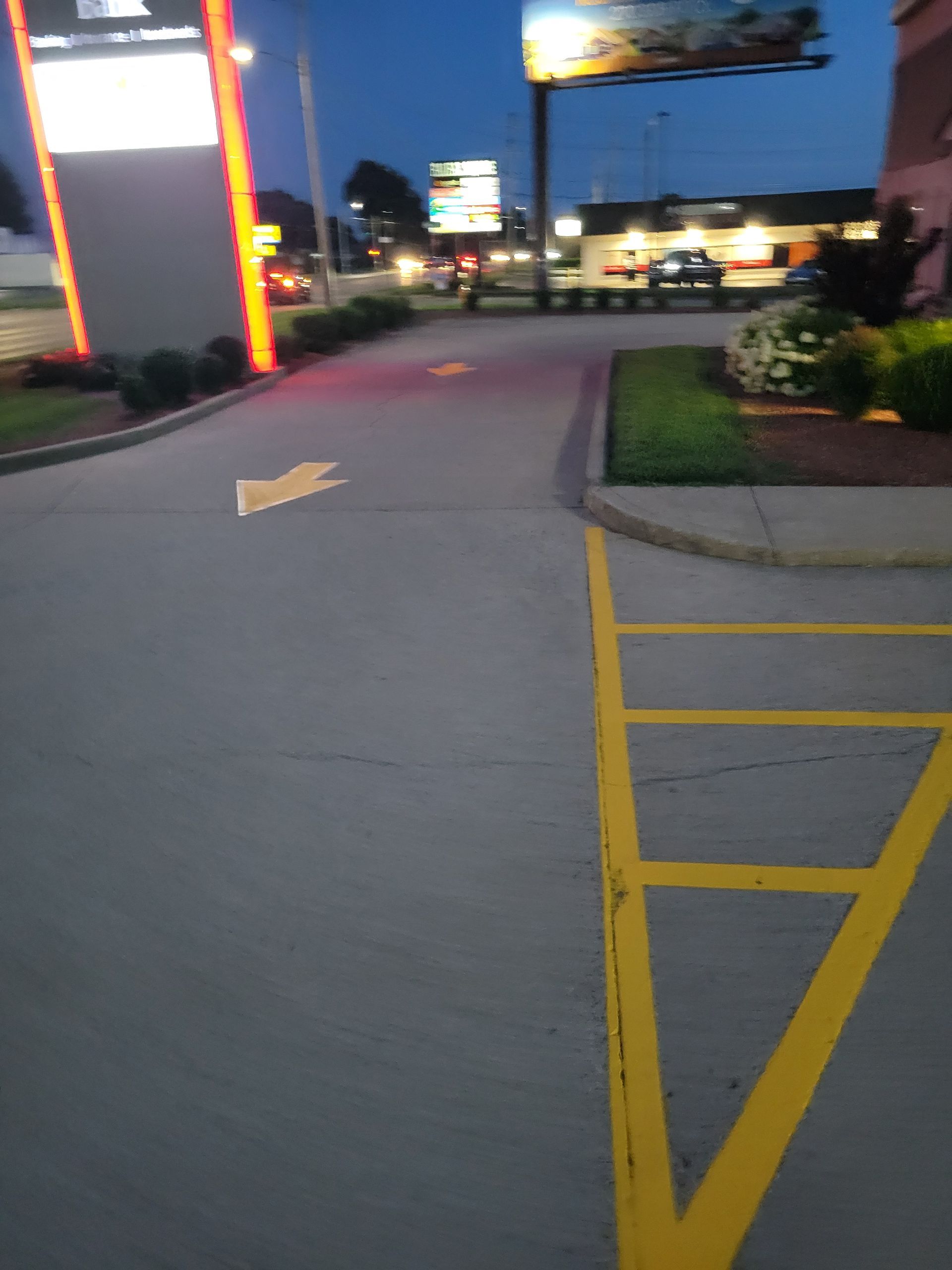 Drive-thru lane with yellow lines and arrow guiding vehicles.  A sign and building are visible in the background at dusk.
