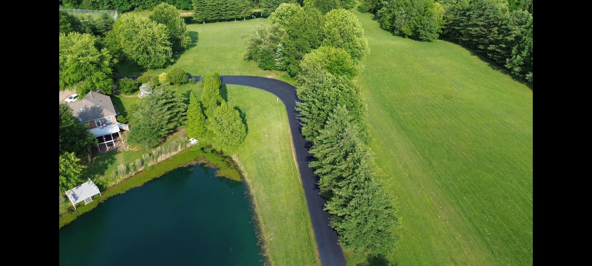 Aerial view of a grassy landscape with a pond, trees, a curving driveway, and a house. The scene is lush green, and sunny.