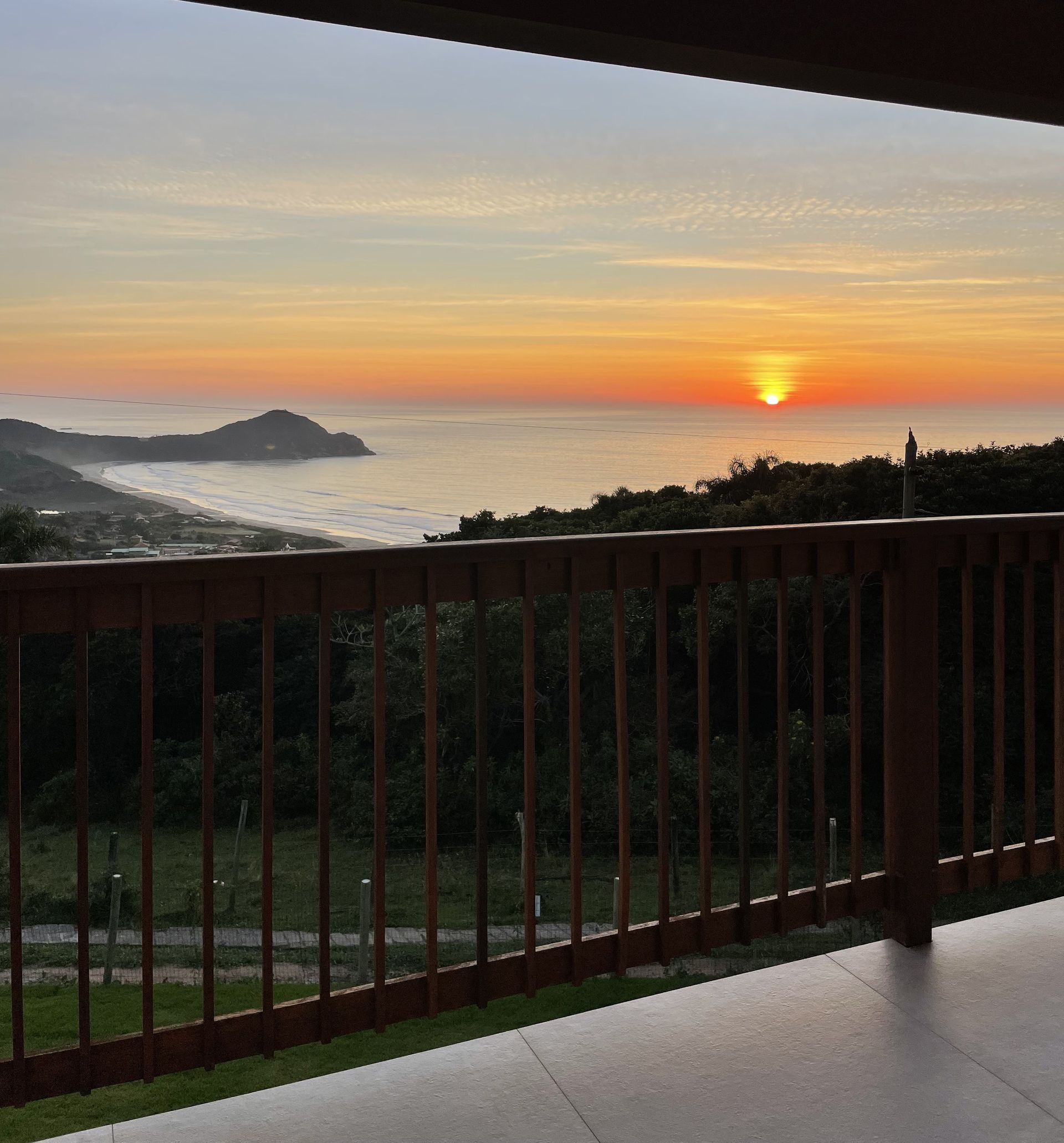 O pôr do sol ilumina o oceano e a costa, vistos de uma varanda de madeira com vista para uma densa linha de árvores.