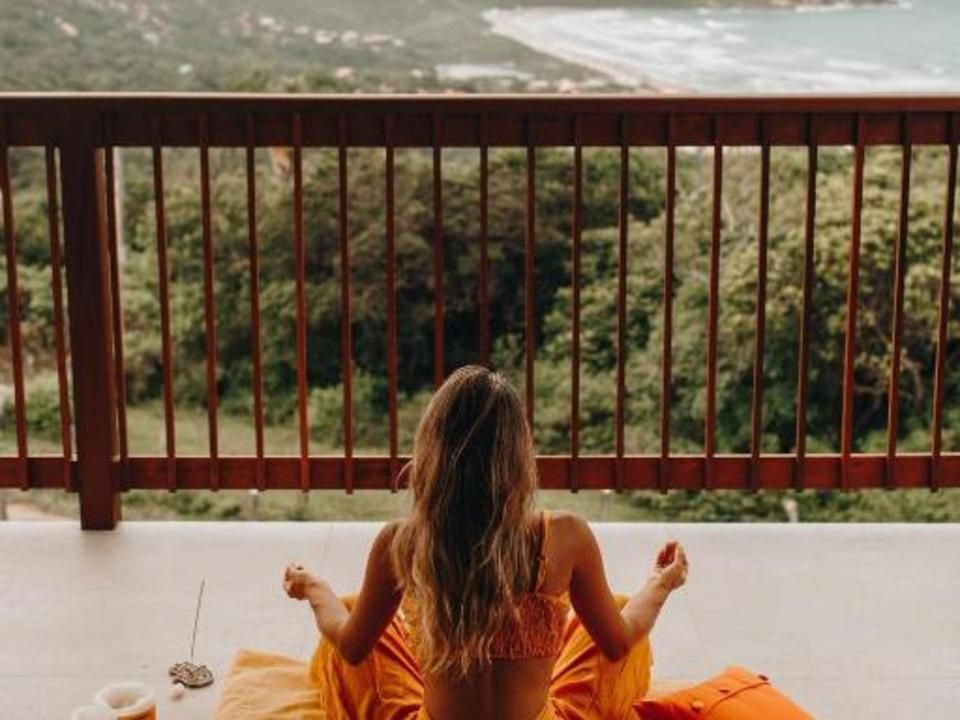 Uma pessoa de cabelos compridos está sentada em posição meditativa sobre uma almofada laranja em uma varanda, com vista para uma floresta e para o oceano.
