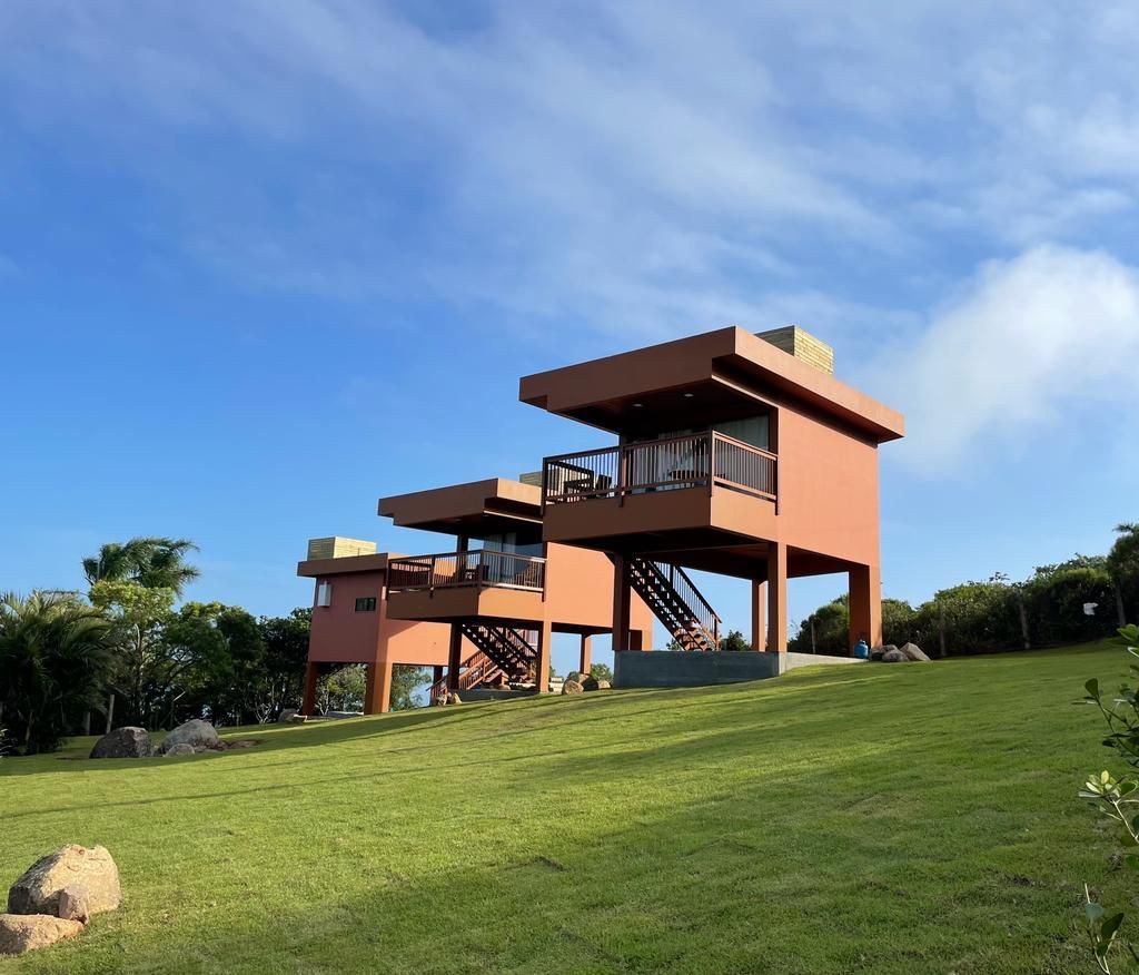 Duas cabanas modernas, elevadas e de cor ferrugem, com varandas, estão situadas em uma colina gramada sob um céu azul brilhante.