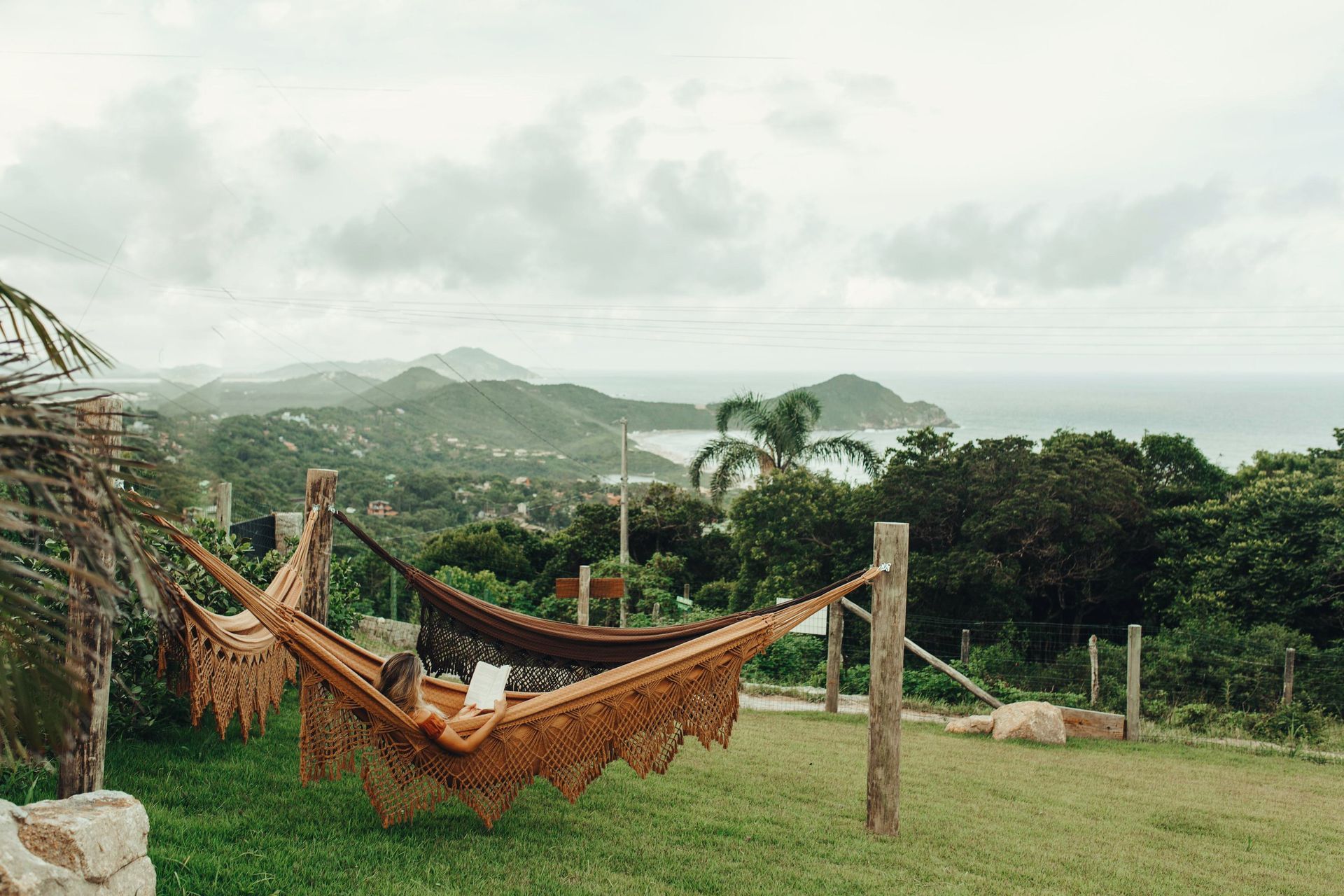 Uma pessoa relaxa em uma rede trançada ao ar livre, com vista para uma paisagem verdejante e exuberante, com colinas distantes e o oceano.