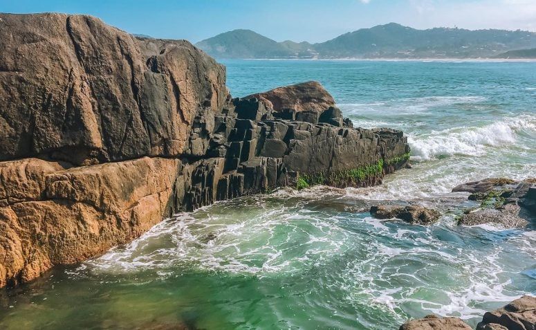 Ondas do oceano quebram contra grandes rochas costeiras texturizadas, com uma piscina natural formada pela maré em primeiro plano, sob um céu claro.