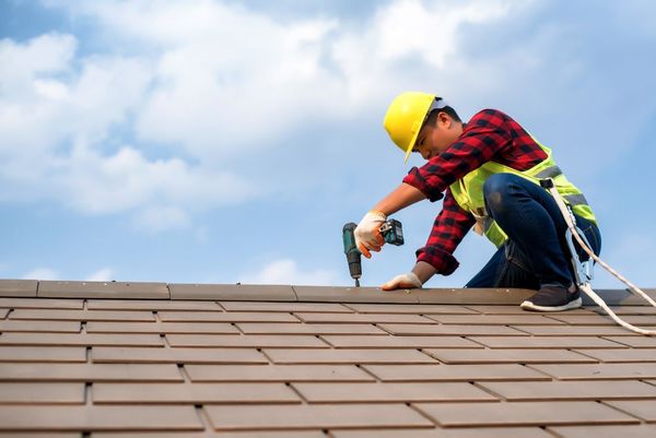 Roofer in hardhat, safety harness, using a power drill on a tiled roof under a cloudy sky.