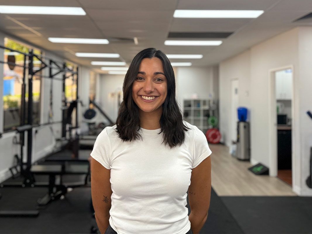Smiling person in a white shirt standing in a gym with exercise equipment in the background