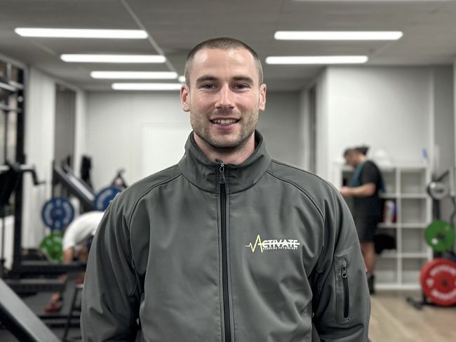 Man in gray jacket standing in a gym, with exercise equipment and people blurred in the background.