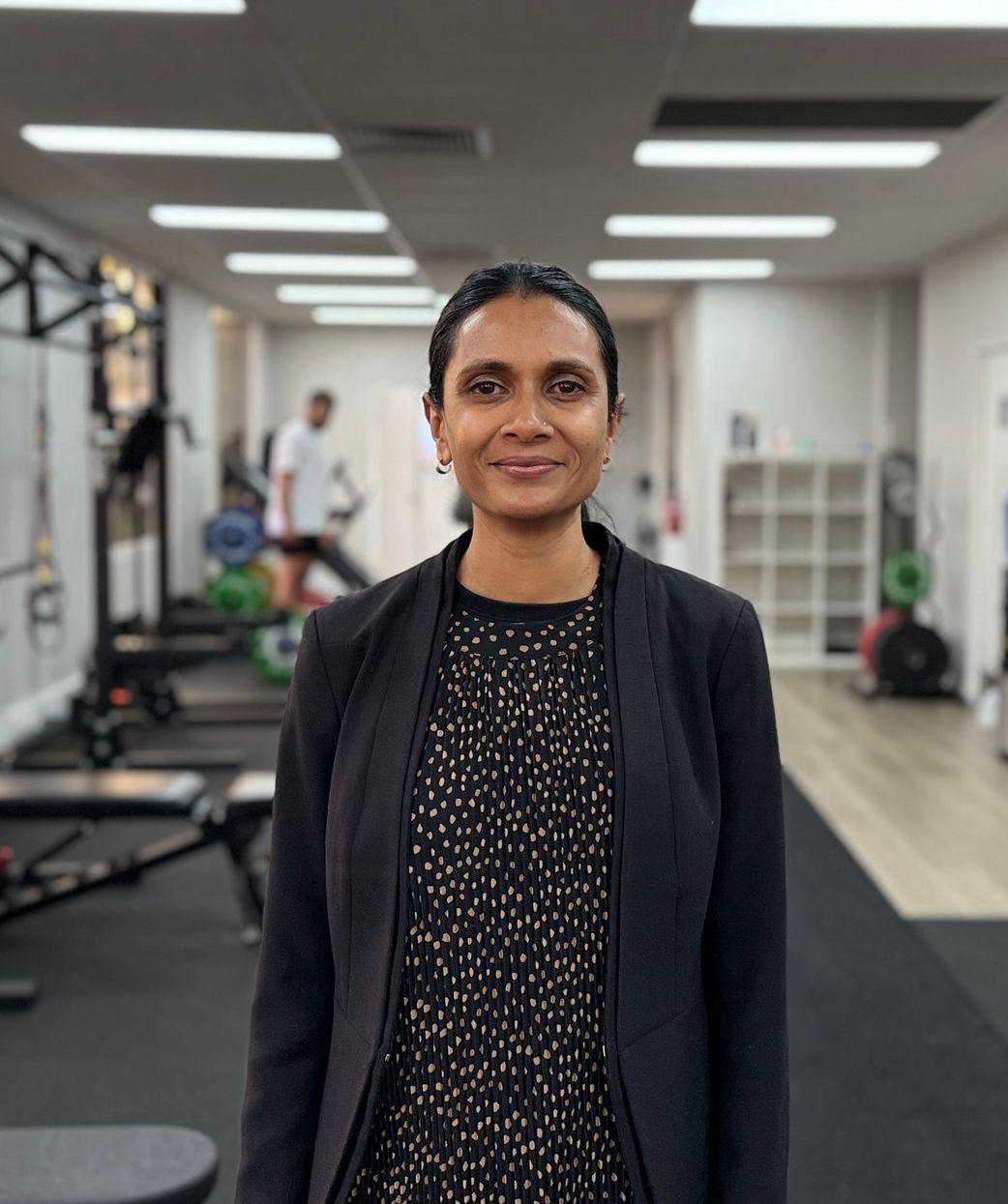 Person standing in a gym, wearing a black jacket and patterned top, with exercise equipment in the background.