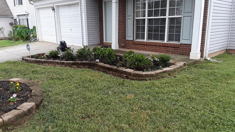A residential front yard featuring a curved brick garden bed with small green shrubs in front of a house.