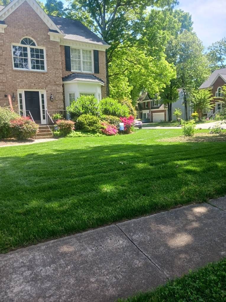 A two-story brick house with a green lawn and blooming landscaping, viewed from a sidewalk on a sunny day.