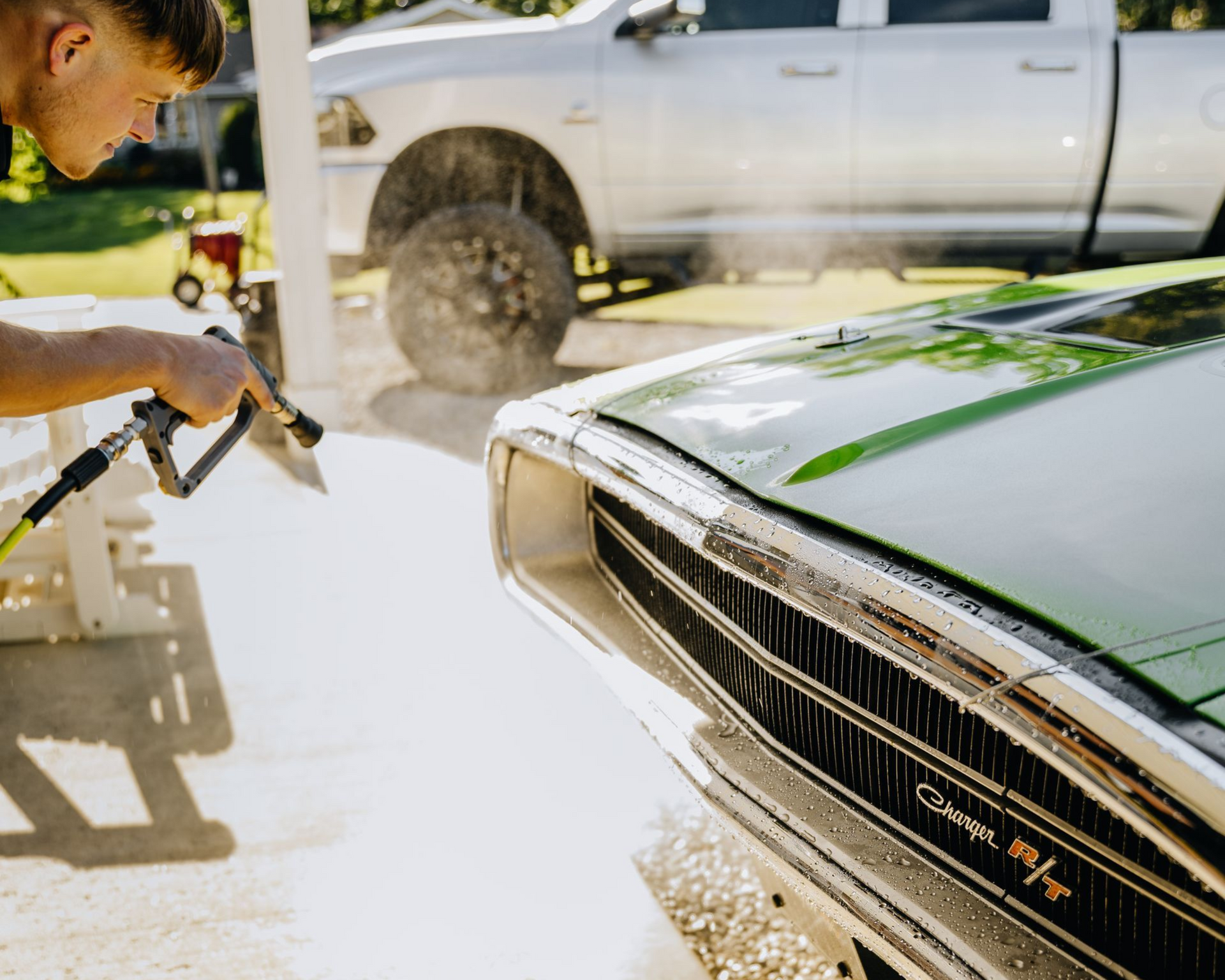Person pressure-washing a green classic car beside a white pickup truck, with water spraying in sunlight
