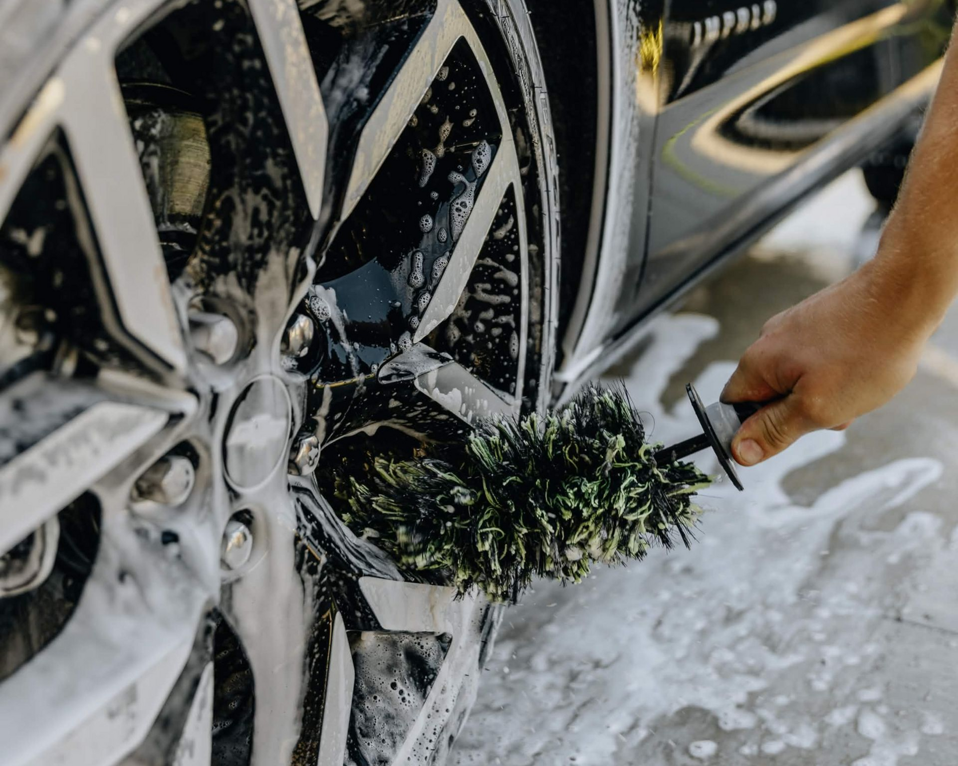 Hand washing a black car wheel with a green brush and soapy foam